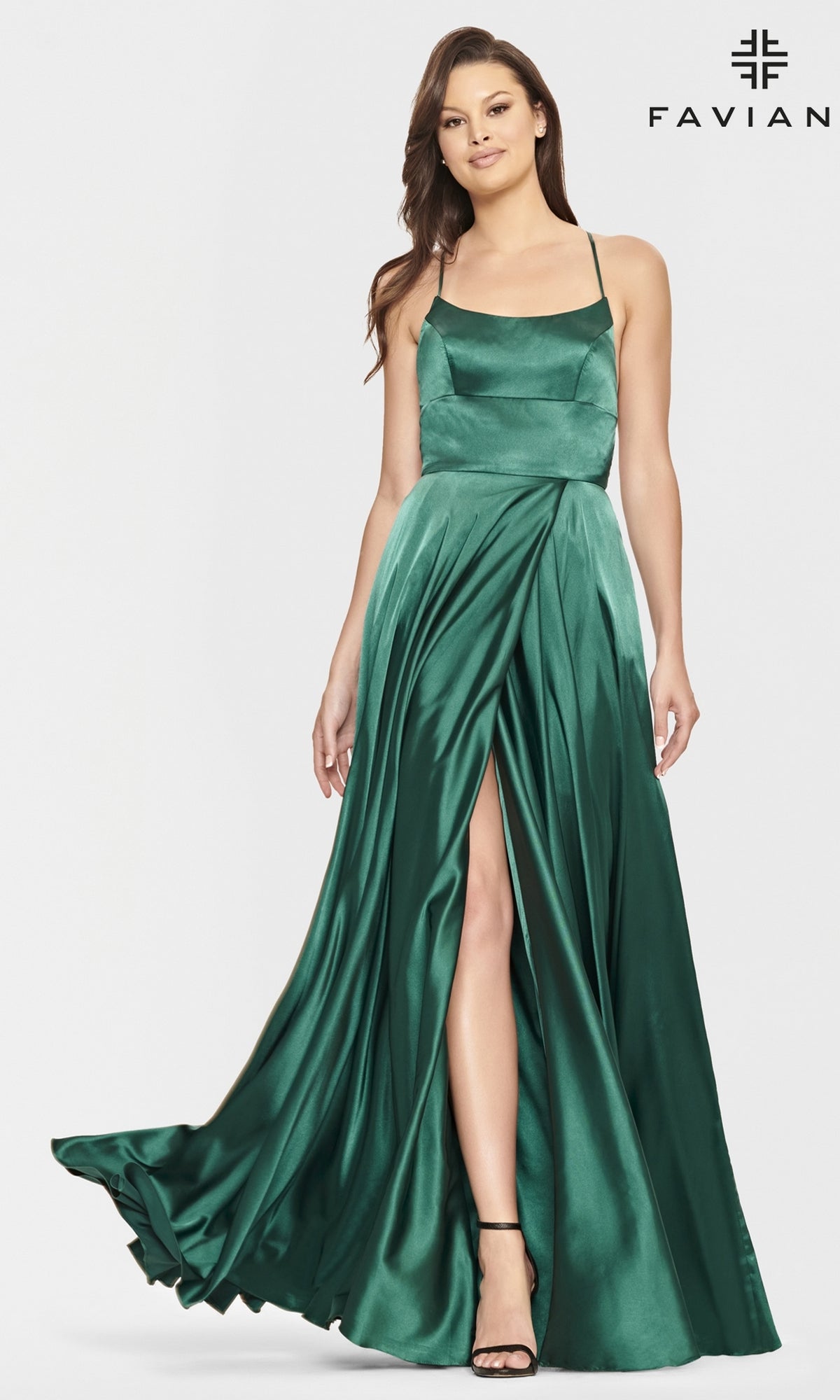 A woman poses against a white background in the Long Open-Back Formal Prom Dress by Faviana—sleeveless, emerald green satin with thin straps and a thigh-high slit. The Faviana logo is visible.